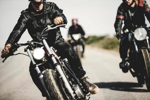 Three men wearing leather jackets riding cafe racer motorcycles along rural Stock Photos
