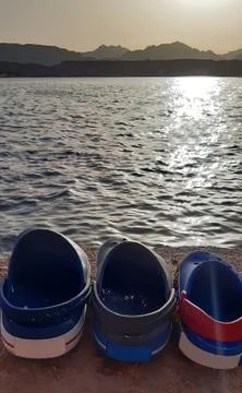 Three multi-colored sandals of different shapes lie in a row on the seashore Stock Photos