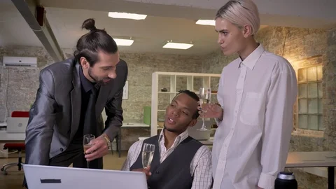 Three multy-ethnic workers tell stories, sitting at laptop in office, drinking Stock Footage 92179823