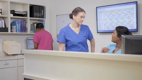 Three nurses at nursing station, two discuss as camera moves left then right Stock Footage 109869851