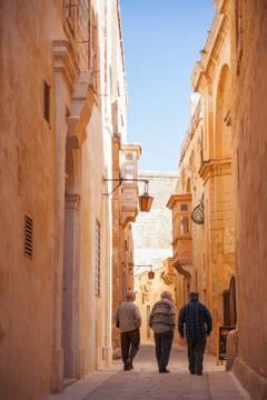 Three old man are walking down ancient narrow street in Mdina, Malta. Stock Photos