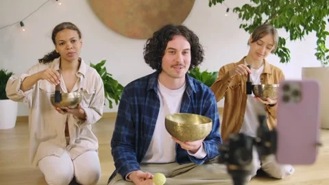 Three people are seated on the floor, using singing bowls and mallets in a sound Stock Footage 346148729