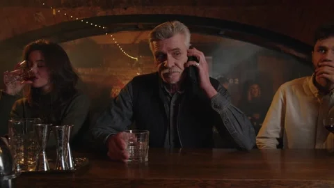 Three people drinks at the bar counter. Adult man answer cellphone while Stock Footage 253630898