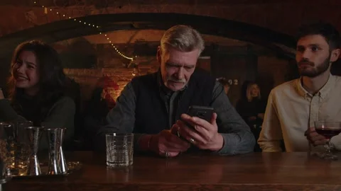 Three people drinks at the bar counter. Adult man checking his smartphone when Stock Footage 253630944