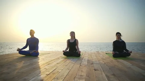 Three persons sitting in the lotus pose on the wooden terrace slow motion Stock Footage