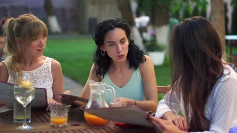 Three pretty girlfriends talking and reading menu in cafe outdoors in the park 스톡 동영상 140675198