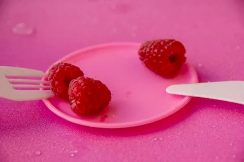 Three raspberry berries lie on a small pink plate, next to a plastic fork and Stock Photos