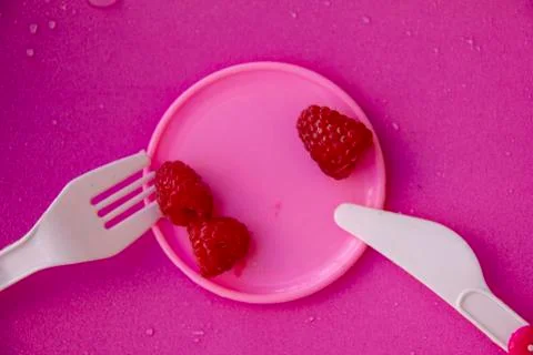 Three raspberry berries lie on a small pink plate, next to a plastic fork and Stock Photos