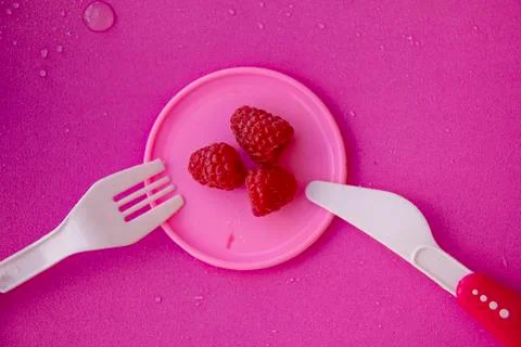 Three raspberry berries lie on a small pink plate, next to a plastic fork and Stock Photos