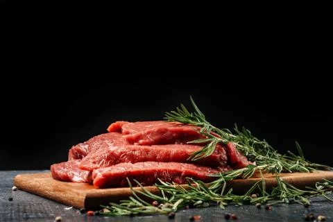 Three raw beef steaks on a cutting board with rosemary on a black background. Stock Photos