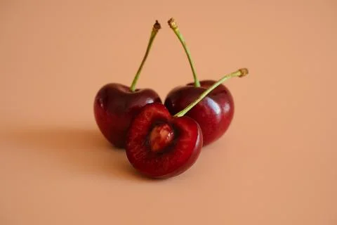 Three red cherries with one cut open on peach background Stock Photos