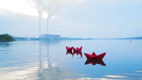 Three red paper boats float on calm water with power plant smoke stacks in .. Video stock 337641161