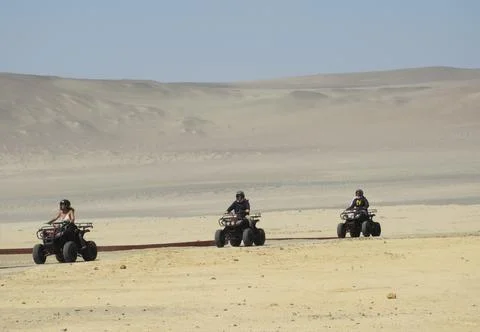 Three riders on quad bikes drive across a vast sandy desert, spaced in a line Stock Photos