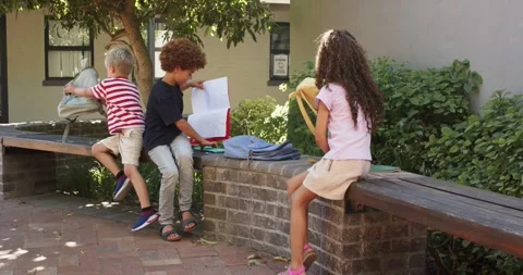 Three schoolchildren sitting on wall at school during break, sorting backpacks, Stock Footage 340980919