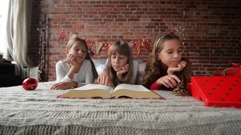 Three Sisters Lie on the Bed in Soft Pajamas, Triplets Read a Huge Book of Stock Footage 96415392