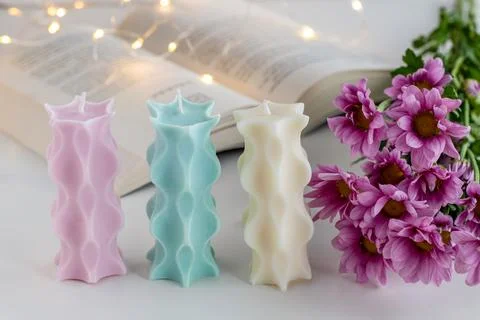Three soy candles on a table with a book, flowers and lights in background. H Stock Photos