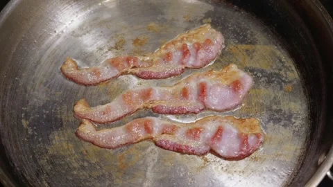 Three strips of bacon sizzle in a stainless steel pan Stock Footage 332748683