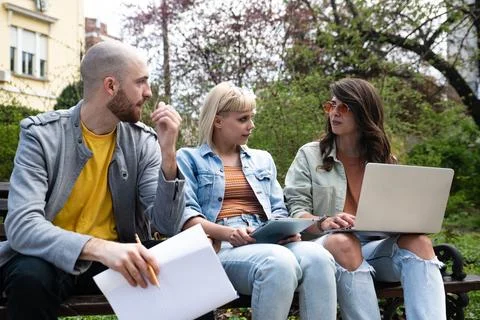 Three students on a bench in the park study talking typing on laptop notebo.. Stock Photos