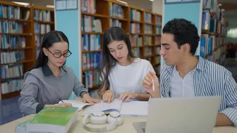 Three students sit together at a library table, studying textbooks with a laptop Stock Photos