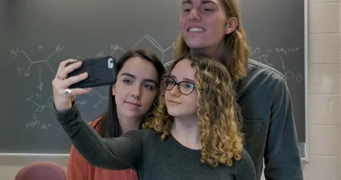 Three students taking selfies in a classroom with a chalkboard in the background Stock Footage 106073967