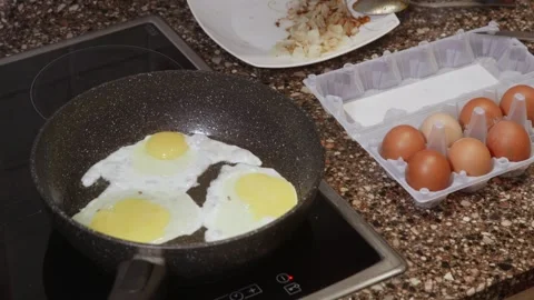 Three sunny side up eggs frying in black pan on stove with egg carton nearby Video stock 323901782
