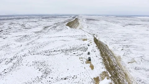Three SUVs moving along the edge of a sandy cliff of the snow-covered desert in Stock Footage 93837432