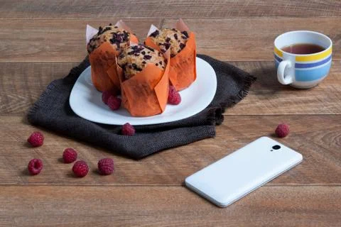 Three tasty muffins with raspberry. Cup of tea and smartphone for communication. Stock Photos
