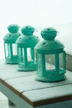 Three teal lanterns with star cutouts glow on a snow-covered table. Stock Photos