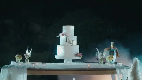 Three Tier White Wedding Cake with Monogram and Flowers on Outdoor Table at Video stock 332920871