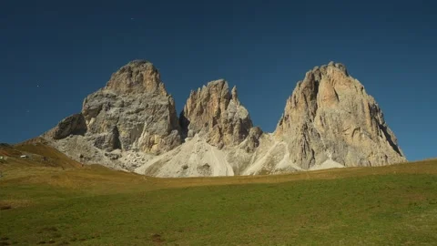 Three towering Dolomite limestone peaks above sunlit alpine meadow under clear Stock Footage 348110038