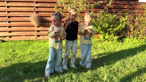 Three triplet sisters blowing soap bubbles in the yard of their house, a fence Stockbeeldmateriaal 281595243