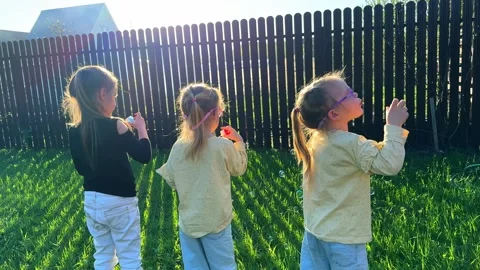 Three triplet sisters blowing soap bubbles in the yard of their house, a fence Stockbeeldmateriaal 281595391