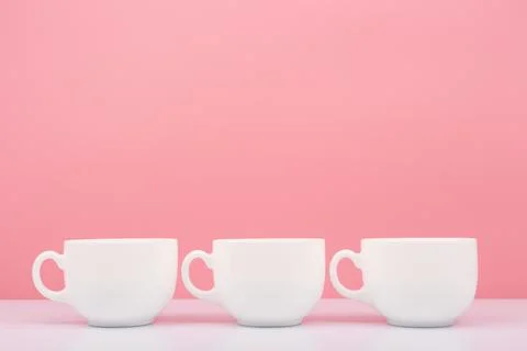 Three white ceramic cups in a row on white table against pink background Stock Photos
