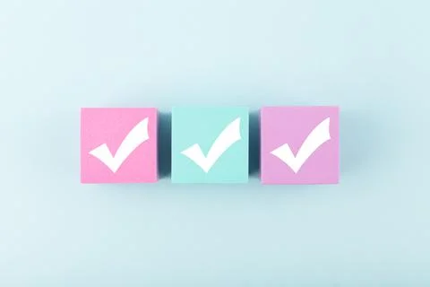 Three white checkmarks on multicolored cubes in a row against bright blue Stock Photos