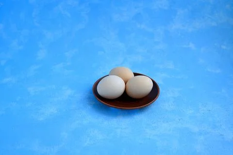 Three White Chicken Eggs on Small Brown Clay Plate with Blue Background Stock Photos