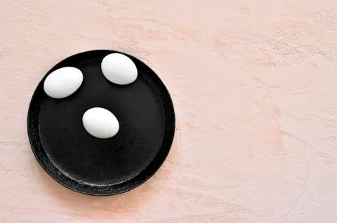Three white eggs in a shell lie on a black pan against the background of a shade Stock Photos