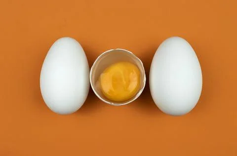 Three White Goose Eggs with One Cracked in the Center on an Orange Background Photos