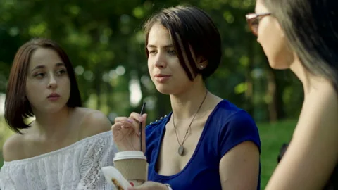 Three women drinking coffee, eating pastries, sitting on grass in a park. 4K Stock Footage 87922053