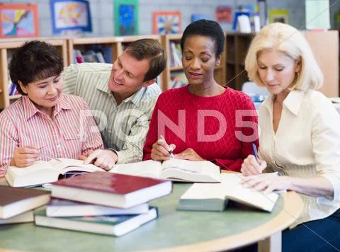 Three women sitting in library with books and notepads while a man leans over Stock Photos