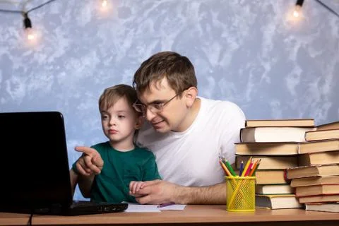 A three-year-old boy with dad is sitting at a desk with books and a laptop. Stock Photos