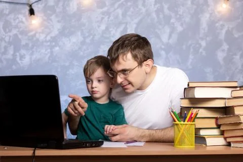 A three-year-old boy with dad is sitting at a desk with books and a laptop. Stock Photos