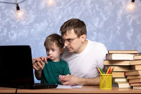 A three-year-old boy with dad is sitting at a desk with books and a laptop Stock Photos
