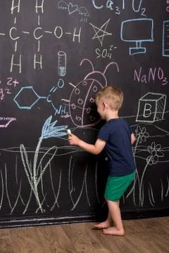 Three-year-old boy draws with multi-colored chalk on a large blackboard. Stock Photos