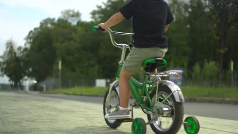Three year old boy rides a small green bicycle on asphalt in the park surrounded Stock Footage 322518302