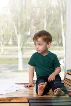 A three-year-old boy sits with a sad look on the windowsill. Stock Photos