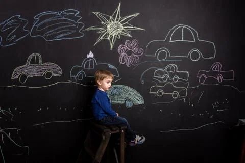 A three-year-old boy sits on a high stool near a slate with different drawings. Stock Photos