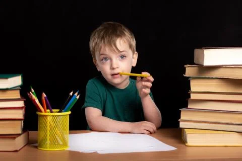 A three-year-old boy sits at a table with books and gnaws a colored pencil. Foto stock