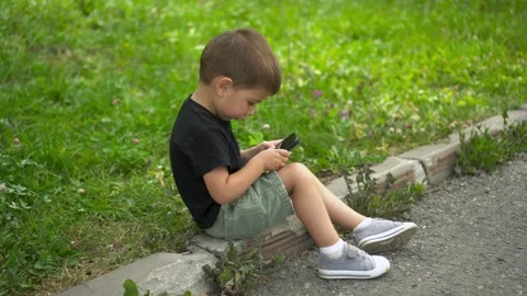 Three year old boy sitting on curb in park playing on smartphone 4k Stock Footage 322518107