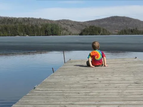 A three year old boy in a tie-dyed t-shirt faces a half-frozen lake from a gr Stock Photos