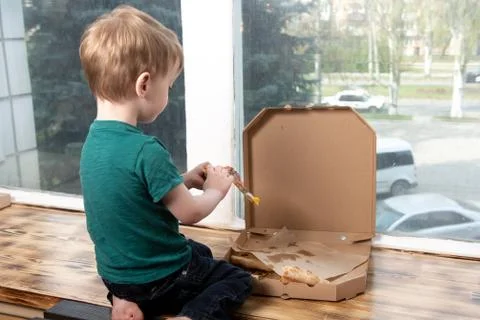 A three year old child eats a slice of pizza from a box. Stock Photos
